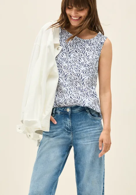 Frau mit gemustertem Top, Jeans und weißer Jacke lächelt.